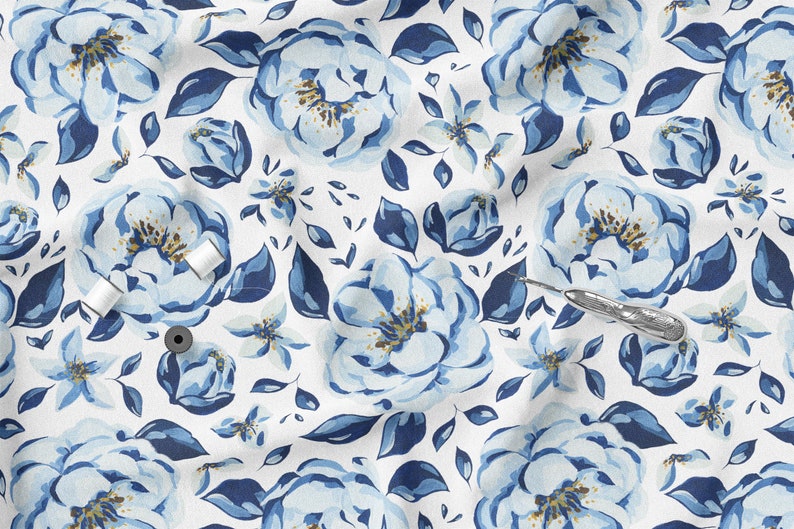 K&ouml;nnte beinhalten: Wei&szlig;er Stoff mit einem sich wiederholenden Muster aus gro&szlig;en blauen Aquarellblumen, kleineren blauen Bl&uuml;ten und dunkelblauen Bl&auml;ttern. Zwei Spulen wei&szlig;en Fadens und ein silberner Nahttrenner sind sichtbar.