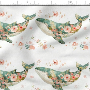 May include: A white fabric with a repeating pattern of teal blue whales covered in pink and green floral designs. The whales are swimming in a sea of pink and green flowers.
