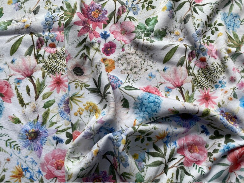 May include: White fabric with a floral print featuring pink, blue, and green flowers. The fabric is folded and draped, showcasing the intricate details of the floral pattern.