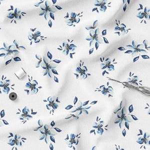 K&ouml;nnte beinhalten: Wei&szlig;er Stoff mit einem sich wiederholenden Muster aus blau-wei&szlig;en Blumenmustern. Das Bild enth&auml;lt drei Spulen wei&szlig;en Fadens, eine schwarze Spule und einen silbernen Nahttrenner. Geeignet f&uuml;r N&auml;h- oder Bastelprojekte.