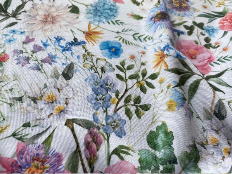 May include: A white fabric with a floral print featuring various flowers in shades of pink, blue, and yellow. The flowers are arranged in a random pattern and are surrounded by green leaves.