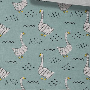May include: A teal blue fabric with a repeating pattern of white geese with yellow beaks and black eyes. The geese are surrounded by black abstract shapes and wavy lines.