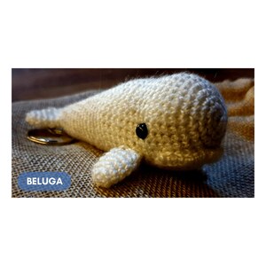 K&ouml;nnte beinhalten: Wei&szlig;er geh&auml;kelter Beluga-Wal-Schl&uuml;sselanh&auml;nger mit schwarzen Augen. Der Schl&uuml;sselanh&auml;nger befindet sich auf einem braunen Jutesack-Hintergrund. Das Wort "BELUGA" ist in Blau auf dem Jutesack geschrieben.
