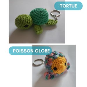 K&ouml;nnte beinhalten: Zwei geh&auml;kelte Schl&uuml;sselanh&auml;nger, einer in Form einer gr&uuml;nen Schildkr&ouml;te mit dem Wort "TORTUE" dar&uuml;ber und der andere in Form eines bunten Fisches mit den Worten "POISSON GLOBE" dar&uuml;ber.