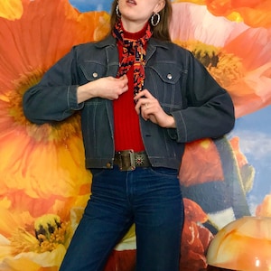 May include: A denim jacket with white stitching, paired with flared jeans. The outfit includes a red turtleneck, a patterned scarf, and a decorative belt. Brown boots complete the look. The background features a floral mural.