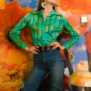 May include: A woman wearing a green and gold patterned long-sleeve shirt with a brown leather belt and brown suede knee-high boots. She is standing in front of a wall with a floral pattern.