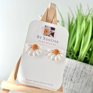 May include: A pair of white daisy stud earrings with textured yellow centers, displayed on a white card labeled BY XIMENA PURE BEAUTY and MADE WITH LOVE IN TEXAS. The earrings are mounted on a small wooden easel stand for clear presentation.