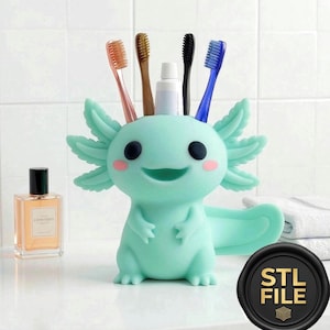 May include: A light teal axolotl-shaped toothbrush holder with a smiling face, holding four toothbrushes and a tube of toothpaste. The holder has pink cheeks and a tail. A perfume bottle and a black and gold "STL FILE" seal are also visible.
