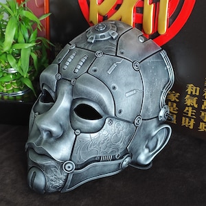 Puede incluir: Una máscara plateada de estilo steampunk con diseño de rostro humano. La máscara presenta intrincados detalles mecánicos, incluyendo remaches y placas. La máscara tiene un acabado metálico liso y está diseñada para cubrir la cara.