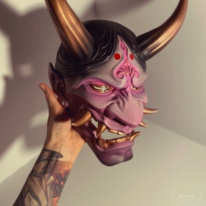 May include: A pink and gold Oni mask with horns, sharp teeth, and intricate details. The mask has a red dot on the forehead and a gold design on the nose.