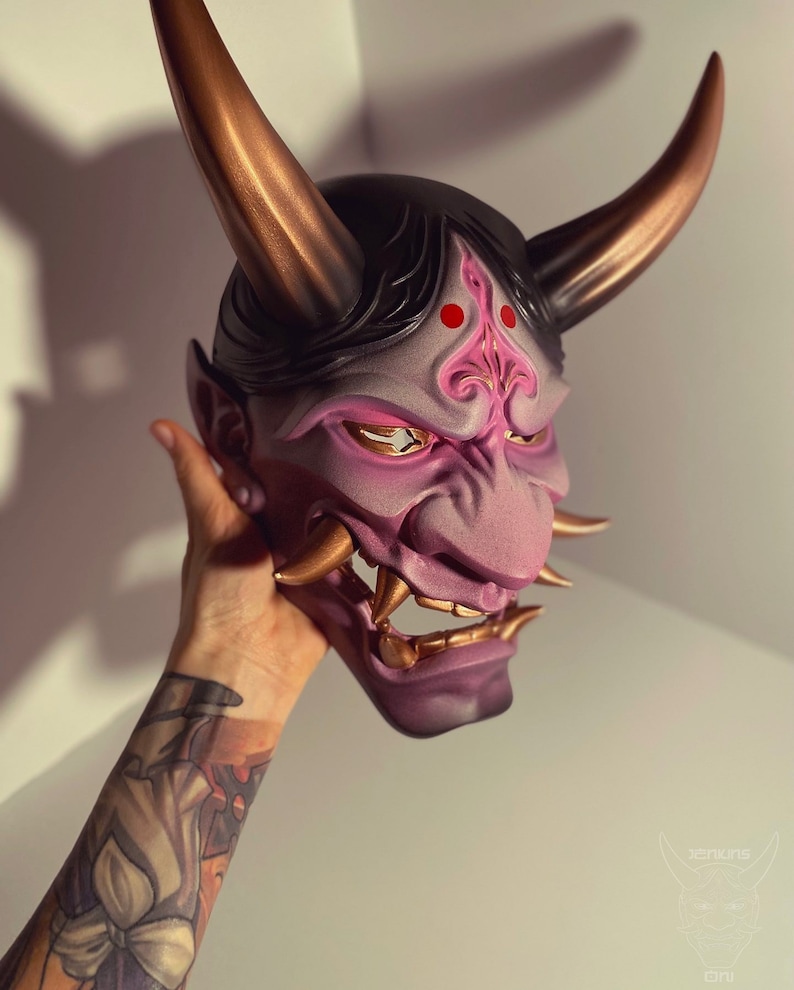 May include: A pink and gold Oni mask with horns, sharp teeth, and intricate details. The mask has a red dot on the forehead and a gold design on the nose.