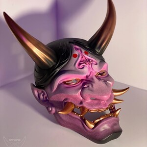 May include: A purple and gold Oni mask with a fierce expression. The mask has sharp teeth, horns, and a detailed design.
