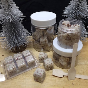May include: A collection of white, cube-shaped exfoliating scrub pieces with red speckles, packaged in two clear plastic jars with white lids and one clear plastic clamshell tray, accompanied by two small wooden scoops.