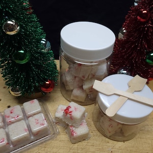 May include: A clear plastic jar with a white lid filled with white, square-shaped wax melts containing red peppermint-like flecks. A plastic tray of identical wax melts and a smaller jar with two wooden scoops are also present.
