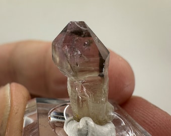 May include: A clear quartz crystal with a smoky purple hue. The crystal is faceted and has a pointed tip. It is mounted on a clear display stand.