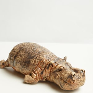 Puede incluir: Figura de hipopótamo de cerámica en tonos marrones y beige. La escultura representa un hipopótamo reclinado con textura detallada y apariencia realista. La figura es un objeto decorativo.