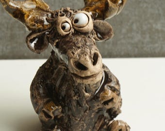 May include: A whimsical ceramic moose figurine with large, googly eyes and detailed antlers. The moose is primarily brown with lighter brown accents on the antlers and a textured surface. The figurine is sitting down, adding to its playful appearance.