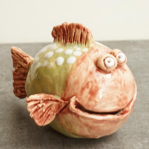 Op de afbeelding: Een grillig keramisch visbeeldje met een lachend gezicht. De vis heeft een groene body met witte stippen en een rood-oranje kop, vinnen en staart. Het beeldje heeft grote, expressieve ogen en is een decoratief item.