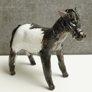 Puede incluir: Figura de caballo de cerámica con pelaje blanco y negro. El caballo tiene una cabeza detallada y está en posición de pie. La figura está sobre una superficie blanca con un fondo gris.