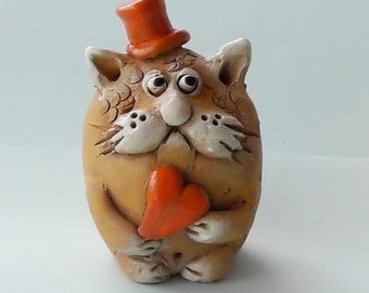Op de afbeelding: Een keramiek kattenbeeldje met een bruin lichaam, wit gezicht en een oranje hoed en hart. De kat houdt het hart in zijn poten.
