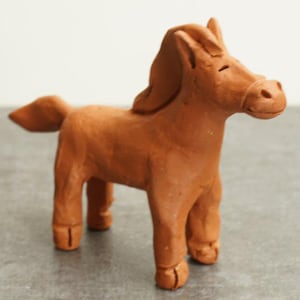 Puede incluir: Pequeña figura de caballo de arcilla hecha a mano en un cálido tono marrón. El caballo está de pie sobre cuatro patas, con una melena fluida y una pequeña cola. La figura es una representación sencilla pero encantadora de un caballo.