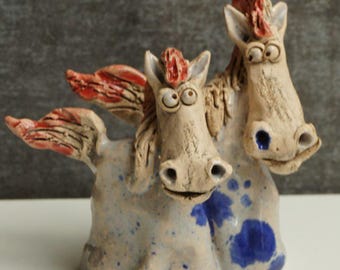 May include: A whimsical ceramic sculpture of two horse-like creatures with red wings and manes. The sculpture has a light beige base with blue and white speckles. The horses have large, expressive eyes and are smiling.