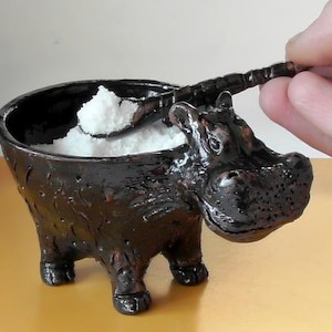 Könnte beinhalten: Eine schwarze Salzschale aus Keramik in Form eines Flusspferdes mit einem kleinen Holzlöffel im Inneren. Die Schale ist mit weißem Salz gefüllt.