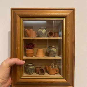 Puede incluir: Una caja de sombra con marco de madera con tres estantes, que muestra macetas, jarrones y una regadera en miniatura de terracota. Algunas macetas contienen flores secas. El marco tiene un borde dorado.