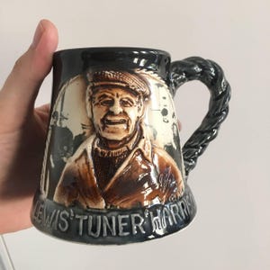 Puede incluir: Una taza de cerámica negra con un asa en forma de cuerda. La taza presenta una imagen en relieve de un hombre con una gorra y barba. El texto "Lewis Tuner Harris" está impreso en la parte inferior de la taza.