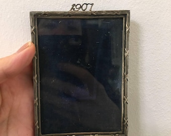 May include: A silver picture frame with a black velvet backing. The frame has a decorative border and the year "1907" is engraved on the top.