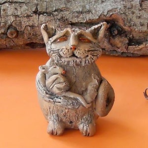以下が含まれることがあります： 猫が小さなネズミを腕に抱いているセラミック製の彫刻。猫は灰色で、表面はざらざらしている。ネズミも灰色で、表面はざらざらしている。彫刻は明るいオレンジ色の背景の上に置かれている。