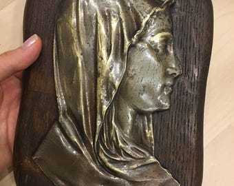 May include: A silver-toned metal relief sculpture of a woman's head, framed in a dark wood frame. The woman is wearing a hooded garment and her head is tilted to the side.