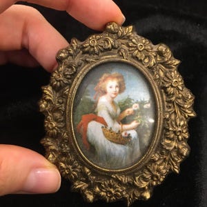 Könnte beinhalten: Ein ovaler Rahmen mit verzierten floralen Details in Goldtönen umrahmt ein Miniaturgemälde einer Frau in einem weißen Kleid, die Blumen hält. Das Gemälde ist vor einem blauen und grünen Hintergrund platziert.
