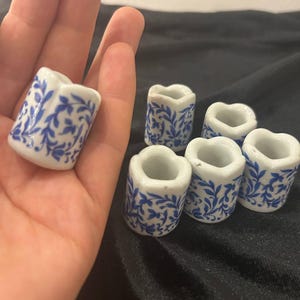 May include: A collection of small, white ceramic holders with a blue floral pattern. One holder is held in a hand, while five others are arranged nearby. The holders have a unique, heart-shaped top.