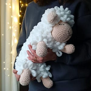 May include: A white crocheted sheep plush toy with pink details. The sheep is lying down with its eyes closed, as if sleeping. The toy is being held by a person.