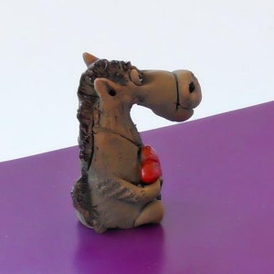 May include: A brown ceramic figurine of a horse holding a red heart in its hooves. The horse has a whimsical expression and is standing on a purple surface.
