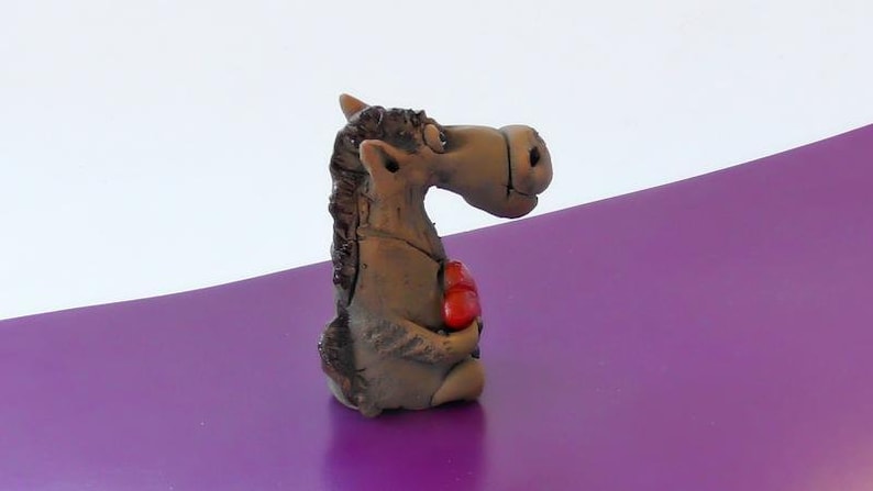 May include: A brown ceramic figurine of a horse holding a red heart in its hooves. The horse has a whimsical expression and is standing on a purple surface.