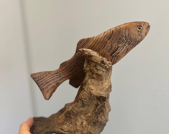 May include: A wooden sculpture of a fish with brown and black stripes, mounted on a piece of driftwood. The fish has a detailed design and a yellow eye. The driftwood is a dark brown color.