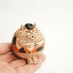 May include: A ceramic cat figurine with a round body, wearing a black hat. The cat has orange eyes and a patterned design in black and orange. The figurine is held in a hand against a white background.