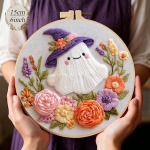 Puede incluir: Arte de aro bordado con un fantasma sonriente que lleva un sombrero de bruja morado adornado con flores. El fantasma está rodeado de coloridas flores bordadas en tonos rosa, naranja, amarillo y morado. El aro mide 15 cm (6 pulgadas) de diámetro.