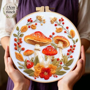 Könnte beinhalten: Gesticktes Hoop-Art mit orangefarbenen und roten Pilzen mit weißen Punkten, umgeben von Herbstlaub und Beeren. Das Kunstwerk hat einen Durchmesser von 15 cm (6 Zoll) und wird auf weißem Stoff vor einem Holzreifen präsentiert.