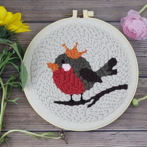 May include: A close-up of a needlepoint hoop with a red, brown, and orange bird wearing a crown. The bird is perched on a branch. The hoop is on a wooden surface with a sunflower and a pink flower.