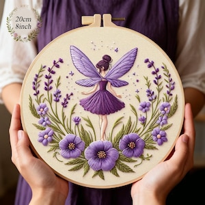 May include: A round embroidery hoop featuring a fairy design. The fairy is dressed in purple and surrounded by purple flowers and greenery. The hoop measures 8 inches in diameter and is held in place by a wooden frame.
