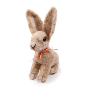 Puede incluir: Una figura de conejo de fieltro hecha a mano en tonos beige y marrón, con orejas largas y una pequeña cinta naranja atada alrededor del cuello. El conejito tiene rasgos faciales detallados y está sentado erguido.