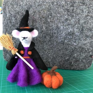 May include: A handmade felt Halloween decoration featuring a white mouse dressed as a witch. The mouse wears a black hat and cape over a purple dress, holding a broom. An orange pumpkin sits nearby. The background is a gray felt board.