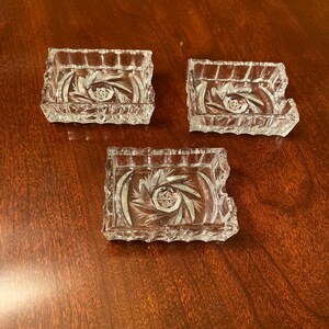May include: Three square clear glass coasters featuring a faceted, starburst-inspired cut pattern on the base. Each coaster has a thick, textured rim with vertical notches, creating a decorative edge that catches the light across the surface.