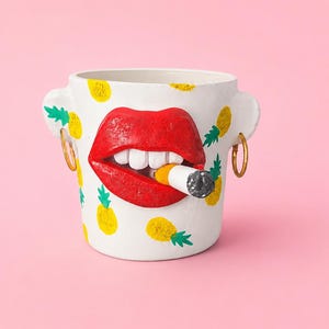 Może przedstawiać: Biała ceramiczna doniczka w kształcie głowy z wystającymi uszami, złotymi kolczykami w kształcie kół i jaskrawoczerwonymi ustami trzymającymi papierosa. Powierzchnia ozdobiona jest wzorami żółtych ananasów z zielonymi liśćmi, wnętrze jest puste.