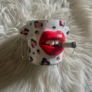 May include: A white ceramic mug with a red lip design and a cigarette in its mouth. The mug features black and pink leopard print spots and gold hoop earrings. The mug is sitting on a white, fluffy surface.