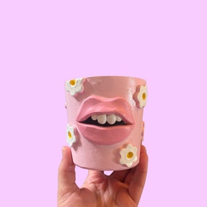 Puede incluir: Maceta de cerámica rosa en forma de labios con flores blancas alrededor del borde.