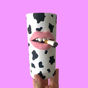 May include: A white ceramic vase with a black and white cow print design. The vase has a pink mouth with white teeth and a cigarette sticking out of it.